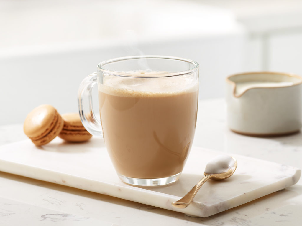 Grande tasse en verre remplie de café au lit, avec 2 macarons et une cuillère à côté.