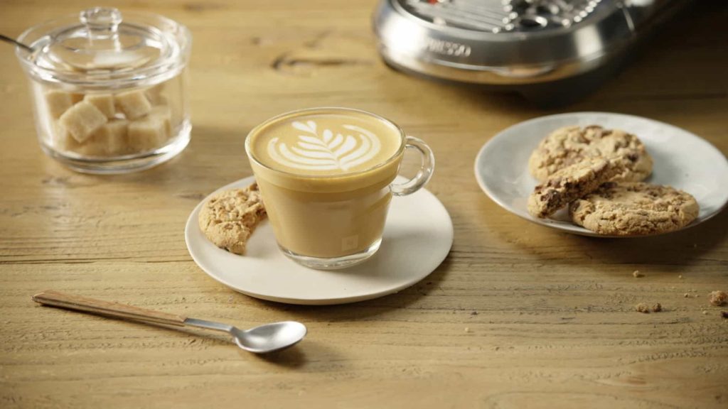 Une tasse en verre de café white flat sur une soucoupe avec à côté du sucre et des cookies.